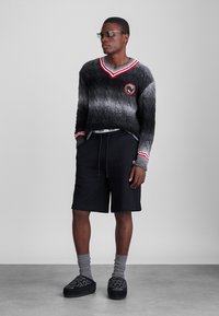 Pull en tricot gris et noir avec des accents rouges et blancs, associé à un short noir, des chaussettes grises et des chaussures noires à motif floral.