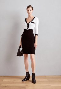 Robe noire et blanche avec manches trois quarts, décolleté en V, détails de boutons, forme ajustée et ceinture décorative. Associée à des bottines noires.