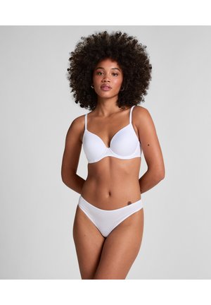 Vrouw met krullend haar, die een witte beha en bijpassende slip draagt, staande tegen een effen lichtgrijze achtergrond, handen achter de rug.