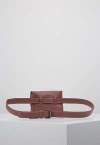 Bolso cruzado de cuero marrón con forma rectangular, correa ajustable y superficie texturizada. Presenta un detalle de hebilla en la parte frontal.