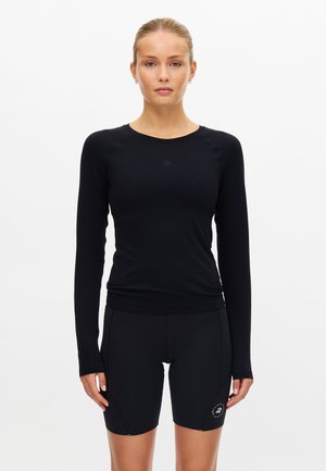 Kvinde stående iført en tætsiddende sort langærmet top og sorte sportsshorts med en lille logo på låret, mod en ensfarvet hvid baggrund.