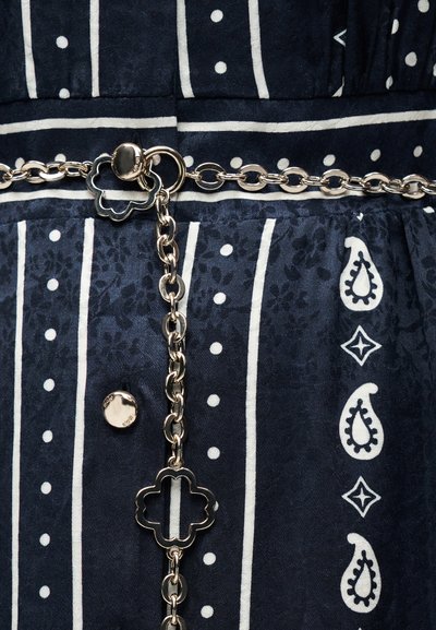 Ασημένια ζώνη αλυσίδα με συνδέσμους σε σχήμα τριφυλλιού, στερεωμένη γύρω από ύφασμα σκούρου μπλε με λευκές ρίγες και σχέδια παϊέτας.