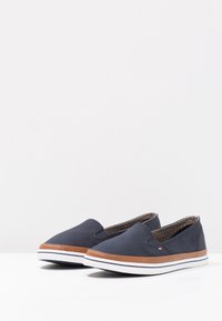 Navy canvas slip-on shoes with brown leather accents, white rubber sole, and minimalistic design. Interior features a patterned lining.