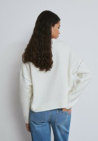 Weißer übergroßer Pullover mit geripptem Saum und Rückendetails mit metallischen Nieten; kombiniert mit hellblauen Jeans.