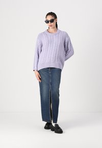 Pull en lilas tricoté avec un design à torsades, associé à une jupe en jean bleu avec une fente frontale. Des chaussures noires et des lunettes de soleil complètent le look.