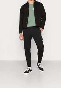 Zwart denimjack, olijfgroen t-shirt met tekst, zwarte cargo joggers en zwarte sneakers met witte zolen. Casual fit met zichtbare zakken.