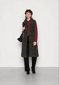 Colete longo, verde escuro, feito de material de felpa texturizado, com dois bolsos frontais, botões e colarinho, usado sobre uma camisa bordô e calças pretas.