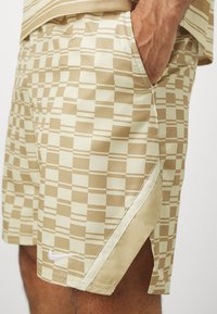 Béžové a krémové kostkované šortky s malým bílým logem Nike, nošené s rukou v pravé kapse, viditelným stehnem a částí pruhovaného trička.