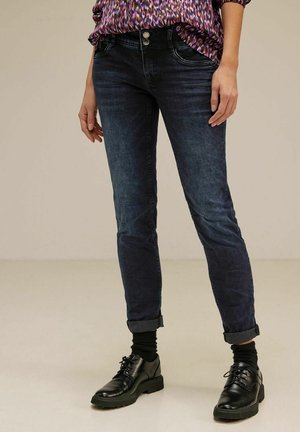 Person iført mørke op-rullede skinny jeans, sorte sokker, sorte snørestøvler og en mønstret top, der står på en ensartet gulv.