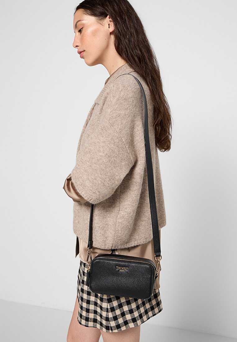 Czarna torba na ramię z zamkiem błyskawicznym i złotymi akcentami. Teksturowany materiał, kompaktowy prostokątny kształt, noszona na długim pasku.