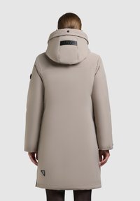 Béžový prešívaný kabát s veľkou kapucňou, bočnými štrbinami a logom naokolo. Hladká textúra, dĺžka po kolená a dlhé rukávy.