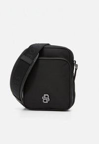 Černá messenger taška s hladkým povrchem, obdélníkovým tvarem a vyraženým logem. Má široký popruh s názvem značky a zapínání na zip.