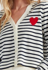 Gestreepte cardigan in wit en marineblauw, met een diepe V-hals, vijf knopen en een opvallend rood vilt hart op de linkerkant van de borst.