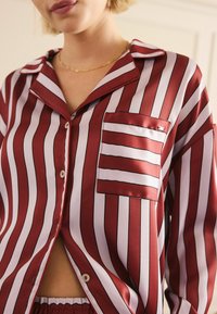 Röd- och vitrandig pyjamasöverdel i slät satin med en bröstficka, notchad krage och knäppning, med kontrasterande svarta konturer.