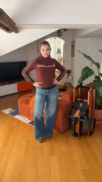 Jeune femme en veste marron et jean se tient dans un salon avec des meubles orange, une télévision, des plantes et un rameur plié.