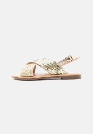 Sandale plate avec des brides tissées métalliques dorées qui se croisent sur les orteils et une bride de cheville avec boucle dorée sur une semelle beige.
