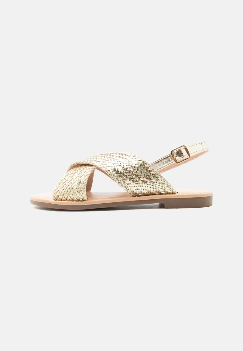 Επίπεδο σανδάλι με μεταλλικές χρυσές πλεγμένες λουρίδες που διασταυρώνονται πάνω από τα δάχτυλα και χρυσή πόρπη στον αστράγαλο σε μπεζ σόλα.