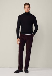 Maglione a collo alto nero, design aderente, abbinato a pantaloni bordeaux, texture liscia. Scarpe nere con dettagli, presentate su uno sfondo neutro.