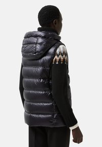 Schwarze Daunenweste mit glänzender Oberfläche, ausgestattet mit einer Kapuze und gestepptem Design. Kombiniert mit einem gemusterten Strickpullover in kontrastierenden Farben.
