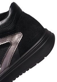 Close-up af en sort sneakerhæl med blandede læder- og ruskindsstrukturer samt en metallisk sølvbroderet logodetalje.