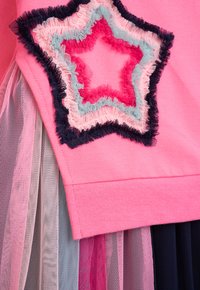 Rosa Oberteil mit einem geschichteten Tüllrock, verziert mit einem bestickten Stern in blauer, rosa und marineblauer Fadenfarbe. Weicher Stoff, kontrastierende Materialien.