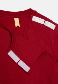 Rød T-shirt lavet af blødt stof med en ribbet halsudskæring, to firkantede velcro-patches på hver skulder og et gult mærke indeni.