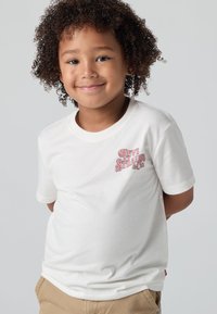 Balta medvilnė marškinėliai su raudonu ir rožiniu Levi Strauss & Co. logotipu kairėje krūtinės pusėje, trumpomis rankovėmis ir apvaliu iškirpimu.