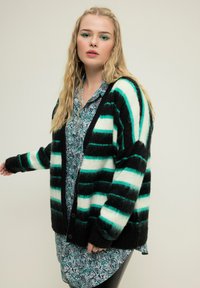 Schwarz-grün gestreifter Cardigan mit flauschiger Textur und Knopfverschluss, über einem Paisley-Print-Kleid getragen. Lange Ärmel.