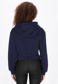 Skrócona granatowa bluza z kapturem wyściełanym podszewką, z prążkowanym dołem i elastycznymi mankietami. Nosi się ją z czarnymi spodniami z wysokim stanem z tylnymi kieszeniami.