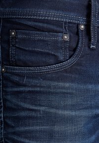 Vaqueros de mezclilla azul oscuro con una textura sutil, detalles en botón plateado y un bolsillo frontal. La tela presenta un ligero desgastado.