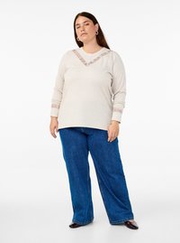 Top beige a maniche lunghe con scollatura a V e bordo in pizzo, abbinato a jeans blu a gamba larga e scarpe nere. Design semplice con accenti testurizzati.