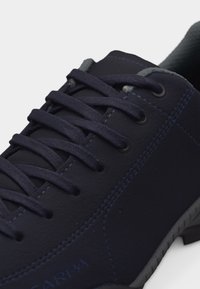 Κοντινό πλάνο αθλητικού παπουτσιού σε ναυτικό μπλε χρώμα με σκούρα κορδόνια, μεταλλικές οπές, εμφανείς ραφές και υφασμένο γκρι περίγραμμα σόλας.