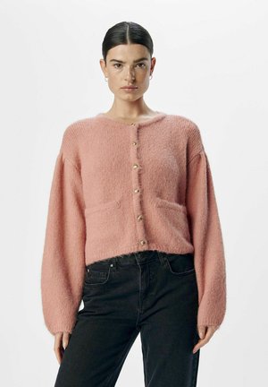 Rosa fluffig kofta med långa ärmar, rund halsringning, framfickor och guldknappar. Bärs över mörka jeans med texturerad yta.