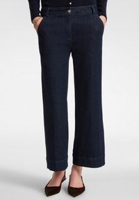 Mujer vistiendo jeans rectos de color azul oscuro con las manos en los bolsillos y zapatos de tacón destalonados de punta marrón sobre un fondo blanco.