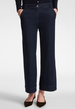 Donna che indossa jeans dritti blu scuro con le mani nelle tasche e scarpe slingback beige a punta su uno sfondo bianco.