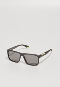 Ochelari de soare dreptunghiulari negri cu lentile închise. Prezintă un finisaj mat, accente verde deschis pe brate și un branding subtil pe ramă.