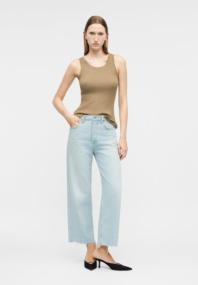 Γυναίκα που στέκεται φορώντας καφέ αμάνικο ριμπ τοπ, ανοιχτό μπλε ξεχαρβαλωμένο τζιν με φαρδιά μπατζάκια και μαύρες ψηλοτάκουνες μυτερές γόβες σε απλό φόντο.