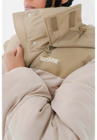 Giacca a vento beige con colletto alto, dotata di logo, bottoni a pressione, chiusura con zip e texture imbottita. Dettagli in Velcro sul colletto.