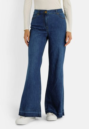 Jeans de mezclilla de corte acampanado en azul oscuro con cintura alta, que cuentan con bolsillos frontales, cierre de botón y zapatillas blancas. Detalle de dobladillo desgastado.