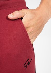 Bordeaux sportieve joggers met een gladde textuur, voorzien van een zijzak en een subtiele zwarte logo bij de zoom. Hand gedeeltelijk in de zak.