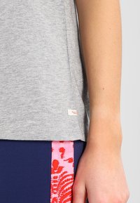 Camiseta de algodón gris con escote redondo; presenta una pequeña etiqueta de la marca en el dobladillo. Combinada con pantalones oscuros con acentos florales en rosa.