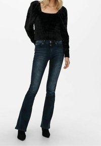 Zwarte vacht top met een vierkante halslijn en puffed mouwen, gecombineerd met donkerblauwe flared jeans. Zwarte enkellaarsjes maken de outfit compleet.