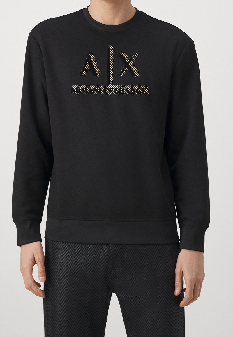 Sweat-shirt noir à manches longues avec logo "A|X Armani Exchange" sur la poitrine, porté avec un pantalon noir à motif chevrons par une personne debout.