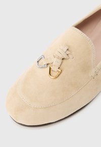 Beige suede loafer with a rounded toe, featuring a knotted lace detail and gold and silver hardware accents. Smooth texture.