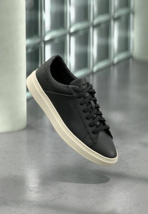 Μαύρο παπούτσι τύπου sneaker χαμηλού ύψους με υφή στο πίσω μέρος και λευκή σόλα, αιωρείται πάνω από λείες σκυρόδεμα κοντά σε παράθυρα με γυάλινα τουβλάκια.