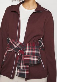 Veste en polaire bordeaux zippée avec un tissu texturé, accents de fermeture éclair blancs, et une chemise écossaise rouge nouée autour de la taille.