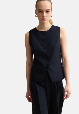 Donna con capelli biondi raccolti in una coda bassa, indossa una canotta nera con bottoni e pantaloni neri larghi coordinati, su uno sfondo chiaro e uniforme.