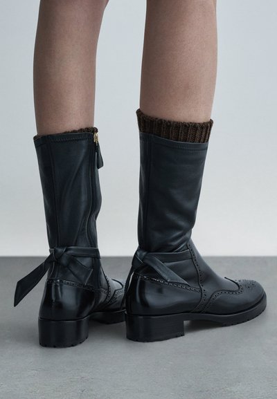 Czarne, skórzane kozaki za kolano z fakturowanym brązowym dzianinowym mankietem, zamkiem błyskawicznym po boku i kokardą z tyłu. Akcenty w postaci wzoru brogue na czubku buta.