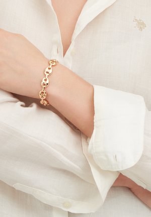 Bracciale a catena d'oro indossato al polso sopra una camicia bianca a maniche lunghe con bottoni e logo ricamato sul petto.