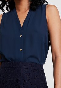 Blusa senza maniche di colore blu navy con scollatura a V, dotata di due bottoni dorati e una tasca discreta, abbinata a una gonna blu navy in pizzo testurizzato.